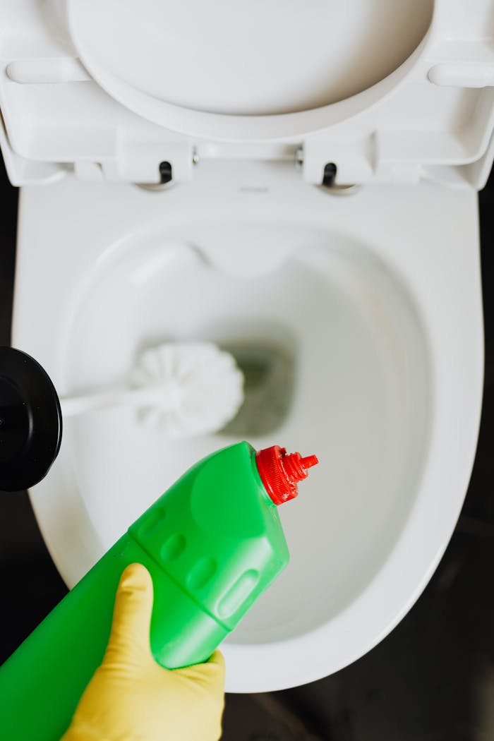 Top view crop anonymous person wearing yellow cleaning gloves cleaning toilet while pouring liquid detergent from green bottle in toilet bowl